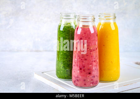 Green, yellow, purple smoothies in bottles of berries, greens, oranges on a white board on a gray table Stock Photo