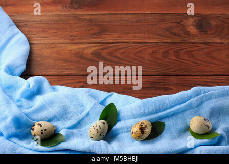 Quail eggs on green leaves on a background of brown wood and blue kitchen cloth. Easter table. Copy space. Stock Photo