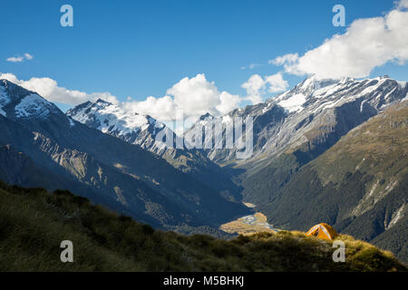 An orange and grey tent on a grassy ridge overlooking a steep valley below the peak of Mount Aspiring