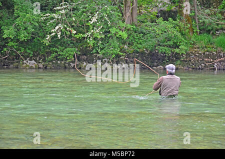 a fly-fisherman casts a fly across a swift river towards the far bank, while wading in deep water Stock Photo