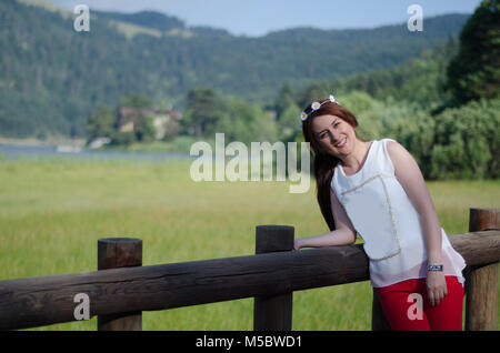 young woman with long hair leaning against wooden handrails, red pants, white shirt, with flower crown in her hair. On a glorious spring day, Stock Photo