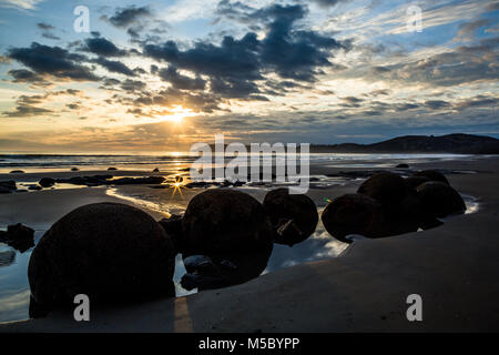 The perfect round boulders of Moeraki Beach in tidal pools at low tide. The sun rises through a cloudy horizon and is reflected in shallow water on th Stock Photo