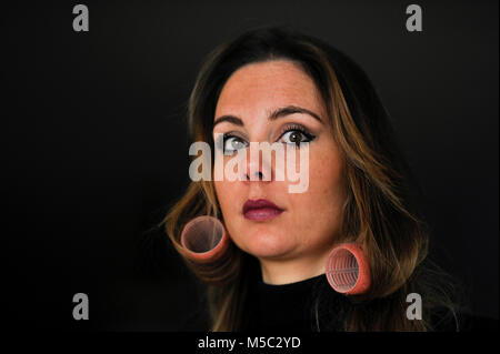 Beautiful young woman with brunette hair wearing hair rollers against dark background Stock Photo