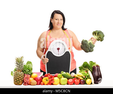 Overweight woman holding a weight scale and a broccoli dumbbell behind a table with fruit and vegetables isolated on white background Stock Photo