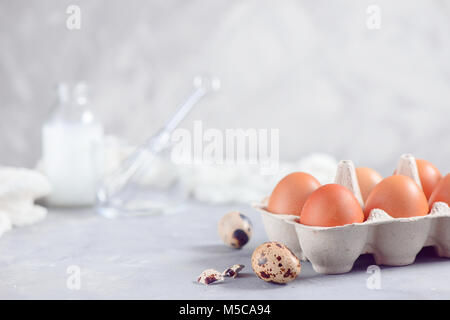 Paper carton of brown eggs on a light background with quail eggs, whisk and ingredients for Easter cooking. High key background with copy space. Stock Photo