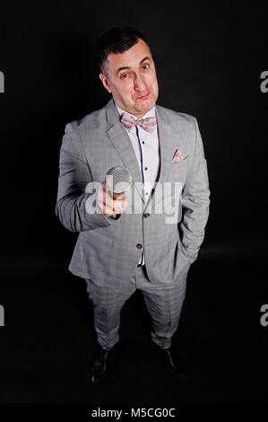 Handsome man in gray suit with microphone against black background on studio. Funny face of toastmaster and showman. Stock Photo