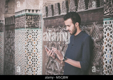 Young Muslim man using his mobile phone in casual clothing beside the wall with traditional Moroccan arabesque decoration Stock Photo