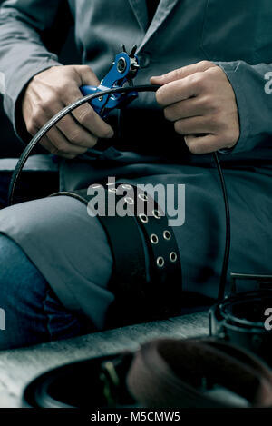 closeup of a young caucasian worker man, wearing a gray working coat, making a hole in a belt with a leather punch Stock Photo