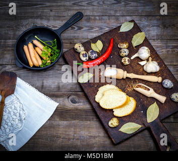 sausages in a black round cast-iron frying pan and raw quail eggs on a gray wooden table, top view Stock Photo