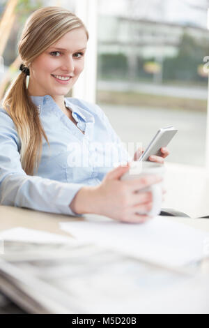 Portrait of confident young businesswoman using mobile phone while holding coffee cup in office Stock Photo
