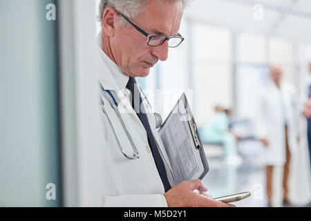 Male doctor with clipboard texting with smart phone in hospital Stock Photo