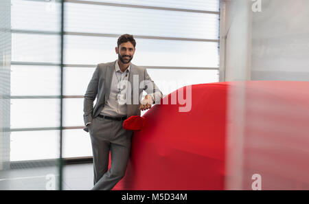 Portrait confident car salesman leaning on covered car in car dealership showroom Stock Photo