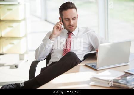 Young businessman using cell phone while reading book at office desk Stock Photo