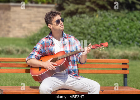 Urban fashionable guy sitting on bench in the park and playing acoustic guitar Stock Photo