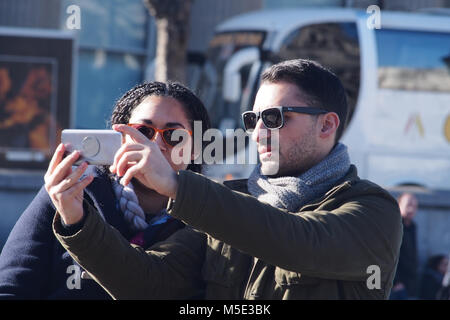 A young couple taking a selfie with a smartphone wearing sunglasses in the winter sunshine in Trafalgar Square, London wearing winter clothing Stock Photo