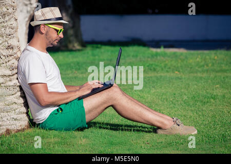 Young freelancer tourist leaning against tree, sitting on grass and working on laptop Stock Photo
