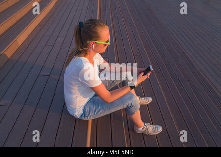 Side view of beautiful modern female siting on stairs, holding smart phone and listening music by using earphones outdoors Stock Photo