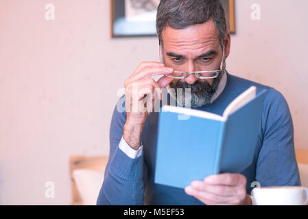 Senior man having troubles reading a book with his glasses, coffee shop surrounding Stock Photo