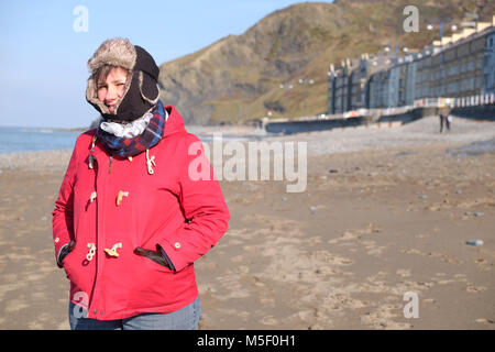 Aberystwyth, Ceredigion, Wales - Friday 23rd February 2018 - UK Weather - Sunny bright weather but cold on the coast at Aberystwyth - A woman walker shows her winter fashion wearing a hat with flaps to wrap up warm whilst walking on the beach. Photo Steven May / Alamy Live News Stock Photo