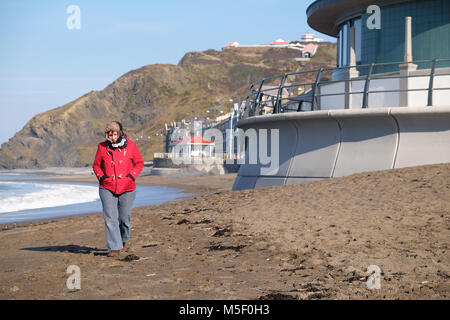 Aberystwyth, Ceredigion, Wales - Friday 23rd February 2018 - UK Weather - Sunny bright weather but cold on the coast at Aberystwyth - A woman walker enjoys a stroll on the beach wearing a hat with flaps to wrap up warm in the cold easterly wind.  - Photo Steven May / Alamy Live News Stock Photo