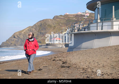Aberystwyth, Ceredigion, Wales - Friday 23rd February 2018 - UK Weather - Sunny bright weather but cold on the coast at Aberystwyth - A woman walker enjoys a stroll on the beach wearing a hat with flaps to wrap up warm in the cold easterly wind.  - Photo Steven May / Alamy Live News Stock Photo