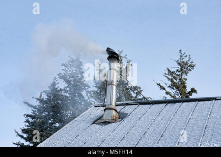 Smoke pouring out of a stove pipe on a metal roof from a wood fire on a winter day. Stock Photo