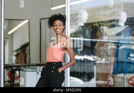 Smiling dress designer in her cloth shop with designer clothes on display. Woman entrepreneur standing at the entrance of her fashion studio. Stock Photo