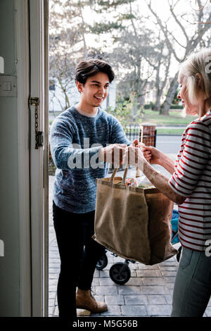 Teenage boy is delivering some groceries to an elderly woman. He is handing her a shopping bag at her front door. Stock Photo