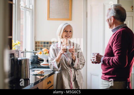 Senior woman is enjoying breakfast and talking to her husband in the kitchen. They vare both drinking tea. Stock Photo