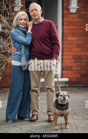 Happy senior couple are posing for the camera outside of their house, with their pet dog on a leash. Stock Photo