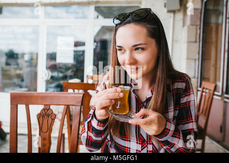 Beautiful young girl with sunglasses on head smiling sitting at table in an outdoor cafe and drinking tea from glass beaker Stock Photo