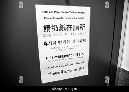 Sign inside a bathroom telling people to throw the paper in the toilet. Written in German, English, Chinese, Japanese, Korean, Arabic and Hindi Stock Photo
