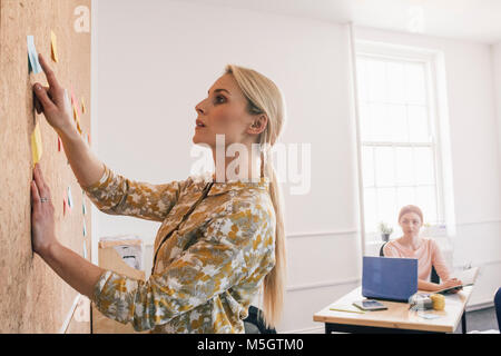 Female worker is pointing to a note on a cork board and is having a discussion with one of her coworkers, who is sitting at a desk in the background. Stock Photo