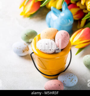 Easter greeting card with colored quail eggs candies in yellow bucket and tulips flowers. Spring holidays concept with copy space. Stock Photo