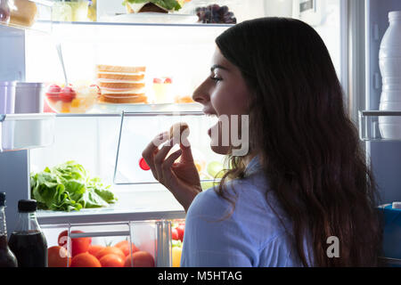 Close-up Of A Woman Eating Cookie Near Refrigerator Full Of Fresh Foods Stock Photo