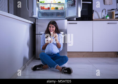 Young Woman Sitting On Floor Eating Slice Of Cake In Kitchen Stock Photo