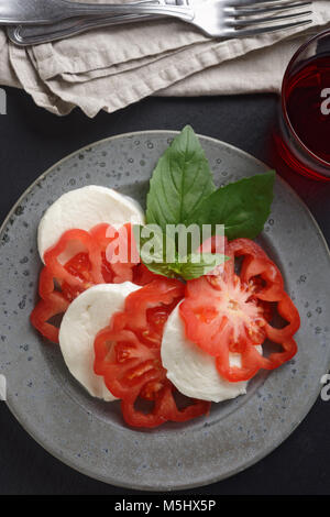 Caprese salad with mozzarella, tomato, and basil leaves and a glass of red wine on a rustic table Stock Photo