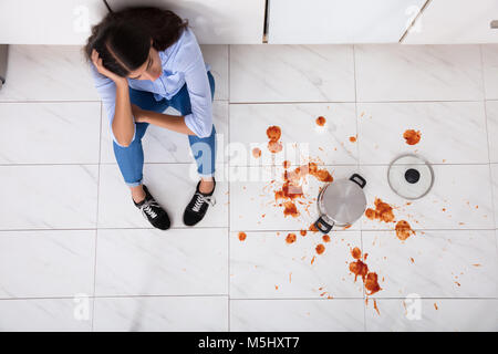 Unhappy Woman Sitting On Kitchen Floor With Spilled Food In Kitchen Stock Photo