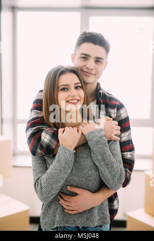 Happy young couple moved to new place to start live together, they are embracing, around many carton boxes with their things. The room is very light a Stock Photo