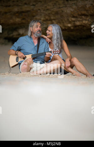 Handsome senior couple sitting on the beach, man playing guitar Stock Photo