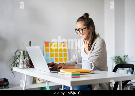 Smiling young woman at home using laptop on desk Stock Photo