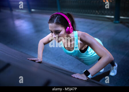 Young woman with pink headphones doing pushups in modern urban setting at night Stock Photo