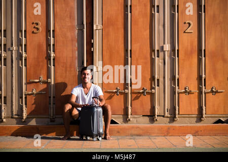 Happy man with rolling suitcase sitting in front of cubicle Stock Photo