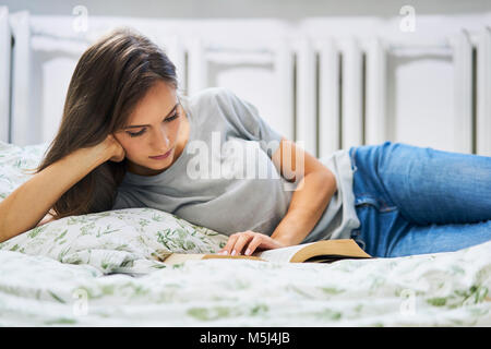 Young woman at home lying in bed reading book Stock Photo