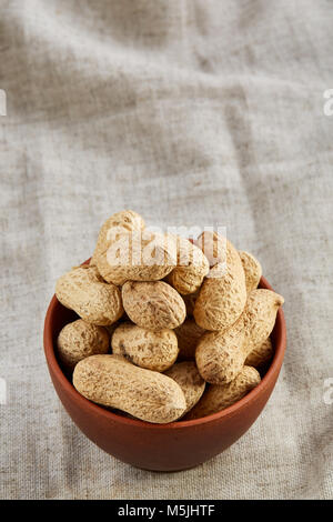Composition of raw unpeeled peanuts in clay ceramic bowl on light grey cotton tablecloth or napkin, some copy space, closeup, selective focus. Healthy Stock Photo