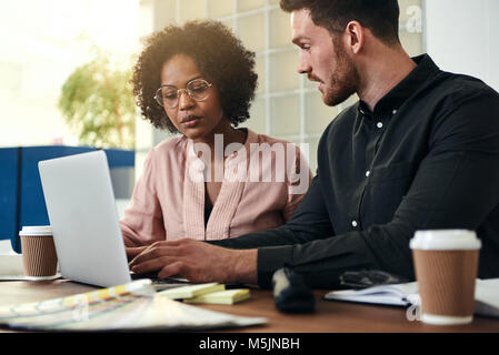 Two work colleagues talking and using a laptop while sitting together at a desk in a modern office Stock Photo