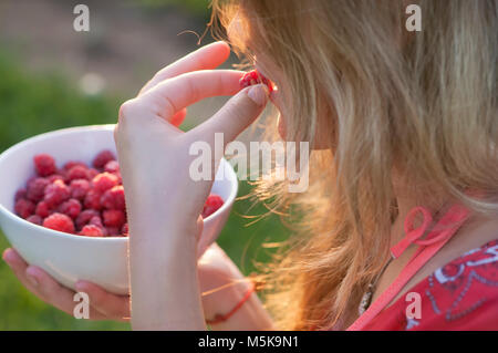 Young girl holding a plate of raspberries, sitting on green grass, summer, dessert, evening, sunset Stock Photo