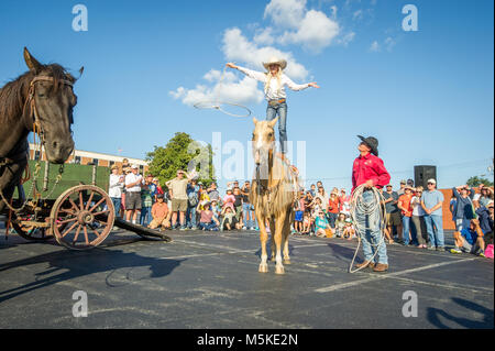 Man in cowboy hat watches young girl standing on top of horse and preforming rope trick with crowd watching, Greensboro, North Carolina. Stock Photo