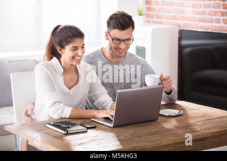 Smiling Young Couple Using Laptop On Table While Having Breakfast At Home Stock Photo