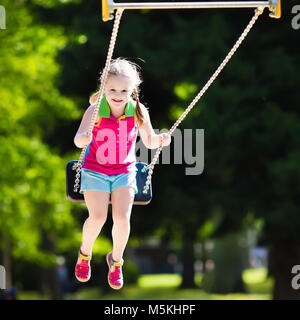 Child playing on outdoor playground in summer. Happy little girl having fun on swing and slide in school yard. Kids play outdoors. Preschooler swingin Stock Photo